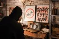 A man in a black hoodie from behind, looking at a laptop and artworks by Banksy.