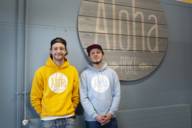 The two founders, young men in yellow and blue hoodies and baseball caps in front of their shop's "Aloha Poke" sign.