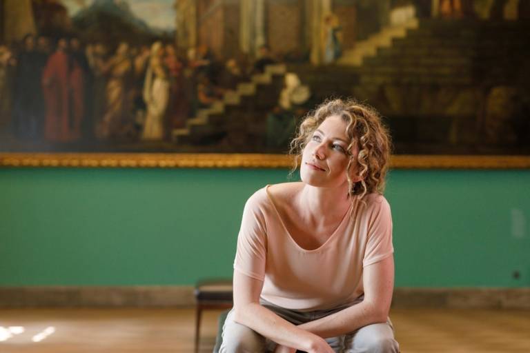 A woman with curly hair is sitting in front of a painting at Sammlung Schack in Munich. 