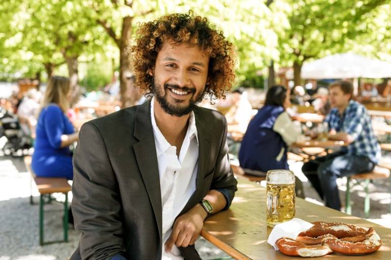 Un homme aux cheveux bouclés et vêtu d'une veste est assis avec des bretzels et de la bière à une table dans un jardin de bière à Munich.